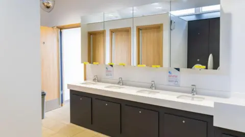 A new block of sinks in a public toilets, with mirrors above, chrome taps, and dark grey cupboards underneath. 