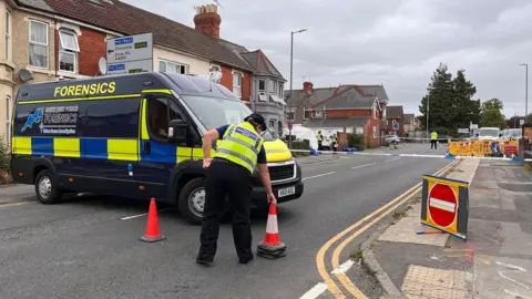 A police officer, seen only from behind, moves a traffic cone in front of a closed road. There is a large police forensics van parked across the road, as well as signs and blue police tape. A white forensics tent can be seen in the distance in front of a house. 