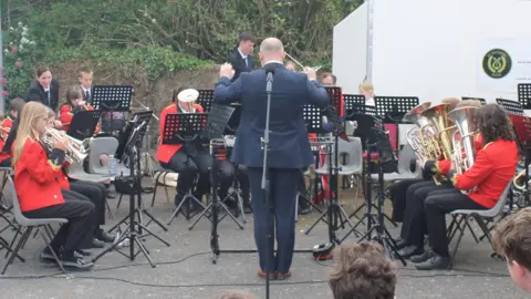 Members of St Keverne Band wearing red jackets and black trousers perform outside with their brass instruments. A conductor wearing a navy blue suit is looking towards the band (and away from the camera) while waving a baton.