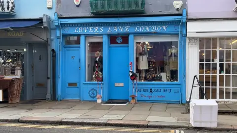 Exterior of a bright blue shopfront labelled “Lovers Lane London”, with large display windows showing clothing and décor and two pink flamingo ornaments standing on the pavement outside.