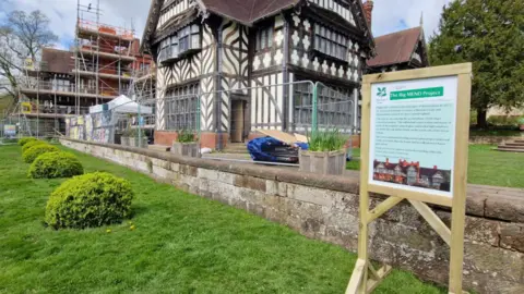 An ornate cream and timber framed 19th Century building, with scaffolding on the outside. A low wall and lawn lies at the front of the building and a sign telling visitors about the Big MEND project is in the foreground.