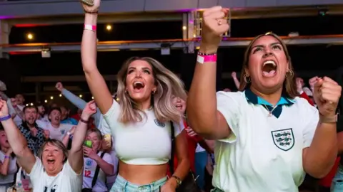 Fans in England football shirts celebrate a goal in a bar. Closest to the camera are two women in white England branded tops and jeans. Behind them and to their right an older woman can be seen celebrating while sat down. Dozens of other fans are in the background. 