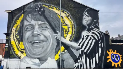 A black and white mural depicts Gary 'Mani' Mounfield, with a close-up of his face in front of a yellow slice of lemon and a larger image of him playing bass guitar in a striped T-shirt