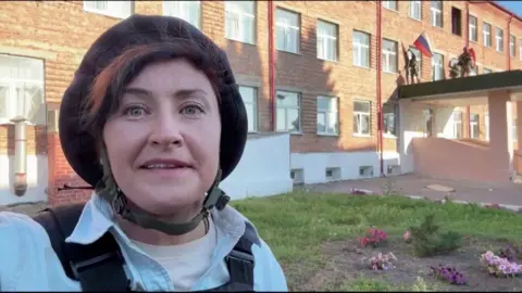 Journalist wearing a protective helmet speaks into the camera arsenic  troops down  her region   a Russian emblem  from a building.