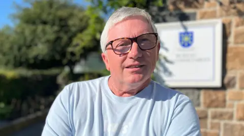 A man with white and grey hair with brown glasses and a blue t shirt. 