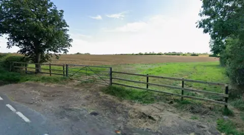 BBC Farmland south of Gartree Road in Great Stretton. A road is in the foreground and trees are either side, inside a fence at the edge of a ploughed field.