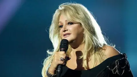 Bonnie Tyler, wearing a black dress, can be seen singing on stage