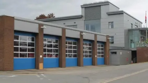 Shropshire Fire & Rescue Service A fire station pictured from the road outside. A high appliance bay can be seen, with four large doors that are blue in between brick pillars. A large grey building is on the right.