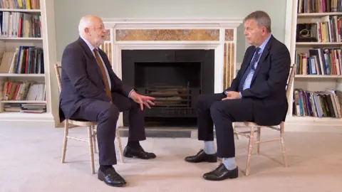 The BBC's Jeremy Bowen sits opposite Philippe Lazzarini with both on wooden chairs. There is a fireplace behind them, which isn't burning, and two white bookshelves filled with books.