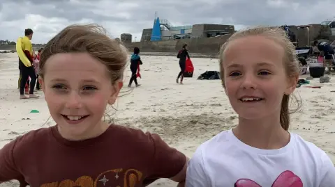 Ella and Jessica standing on a beach. The young girls are standing next to each other and are smiling at the camera.