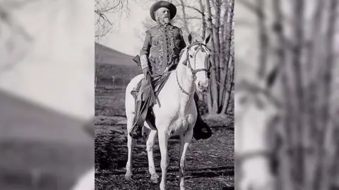 Buffalo Bill Center of the West Sitting on a white horse, an older Cody is pictured with white hair wearing and ornately decorated jacket and cowboy hat, carrying a gun in his right hand 