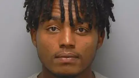 A man with his hair in short locks is pictured facing the camera in a police mugshot. 