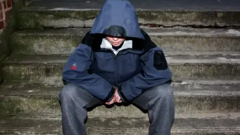 A young boy sits on some steps with a hood up, looking at the floor. He has jeans on and his face is partially hidden.