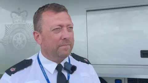 Annabel Amos/BBC Supt Pete Basham is looking towards the right of the camera. He has short brown hair and stubble, and is wearing a white shirt with a tie that has police badges on the shoulders.