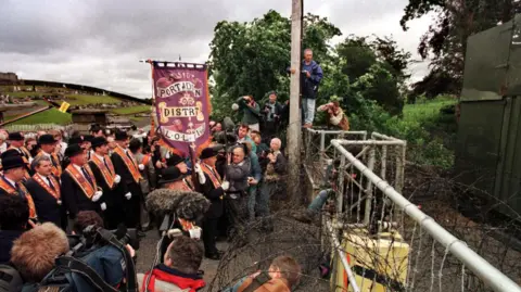 Pacemaker A group of Orange Order members standing at a fence in the middle of a road. 