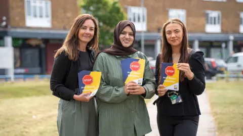 City of Wolverhampton Council Three women are looking at the camera and holding leaflets including the words stop anti-social behaviour. Grass behind them and a building and vehicles further back are blurred.