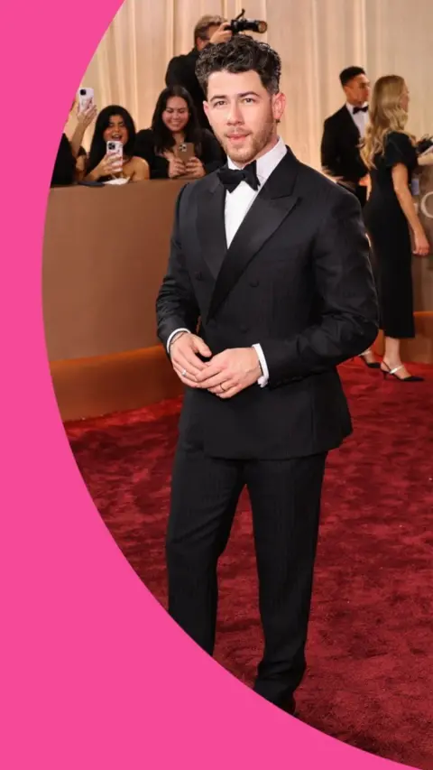 A man, Nick Jonas, stands in a suit on the red carpet