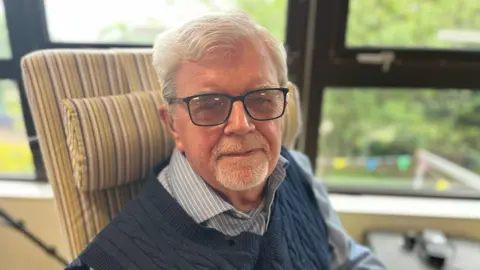 Kevin is looking at the camera and smiling. He is wearing a blue and white stripped shirt and a navy knitted tank top. He has short white hair and glasses.