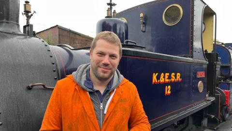 A man wearing orange overalls stood next to a dark blue steam train. The man is smiling, he has blonde hair. The steam engine has yellow letting on the side which reads K.&E.S.R.