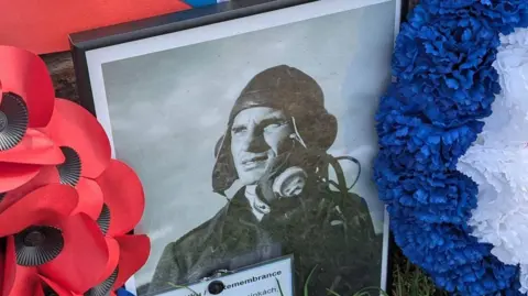 Gary Hadden/@garyh_creates A close-up framed picture of Czech pilot Alois Vasatko placed on the side a memorial made in his memory. In it, he is wearing a uniform and a flying helmet. There is a poppy wreaths and tributes next to it.