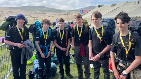 A group of boys wearing walking gear and medals around their neck. They are all looking towards the camera. Tents and the moors are visible behind them