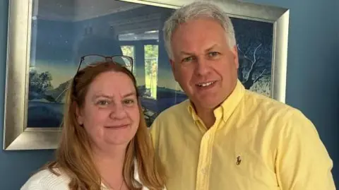 A woman with long blonde hair wearing a white jacket and white top stands next to a man with short grey hair in a yellow shirt.