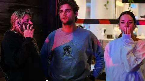 Getty Images A group of concerned onlookers at the scene of the Bondi beach shooting. It's nighttime and the three people's faces are partially lit with emergency service lights.