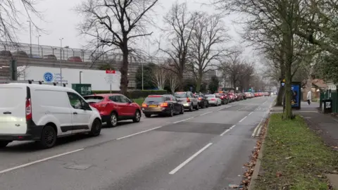 A queue of congested traffic stretches along Wigginton Road heading out of York. In the background is the hospital's multi-story car park, queues for which have been blamed for the regular build up of traffic. The road is lined with bare trees and there is a muddy strip of grass next to the pavement on the opposite side of the hospital.