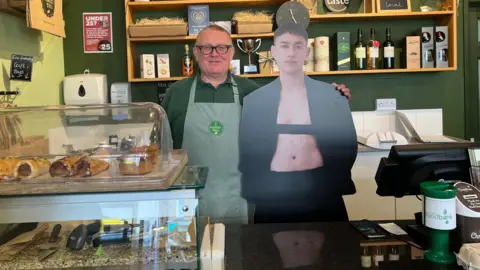 A man at Forest Deli behind the counter with his arm around a cardboard cutout of Olly Alexander