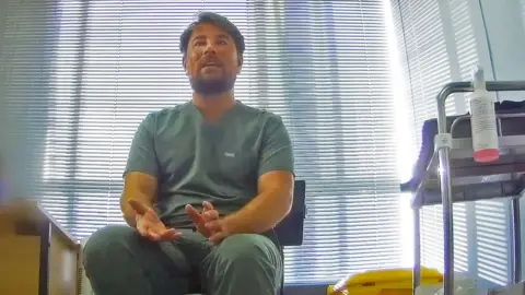 Ricky Sawyer, filmed undercover in his small pop-up clinic at a London office, sits next to a metal trolley wearing green uniform. He is a man in his 20s with straight brown hair, a moustache and beard.