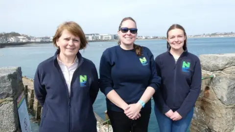 Nature Commission A team photo of Head of Operations and Education Angela Salmon (left), CEO Jessi Jennings (middle) and Ecologist Charlotte Burgoine (right) stood in front of blue sea. There are walls either side of them.