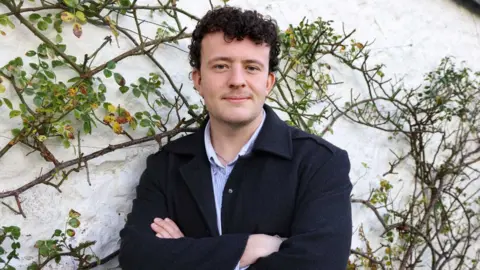 An image of a man in a black suit jacket in front of some greenery