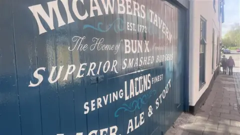 Neve Gordon-Farleigh/BBC The outside fence of the Micawbers Tavern in Norwich. It is painted blue and says, MICAWBERS TAVERN EST 1772. The Home of BUN X... SUPERIOR SMASHED BURGERS. SERVING LACONS FINEST...