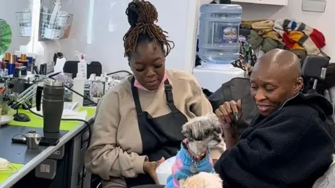Shea Osei Shea Osei (left) wearing an apron. She is working on the nails of Cynthia Erivo in who is sitting in a make-up chair, having her nails done, with a small dog on her lap.