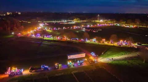A drone shot of brightly-lit trucks and tractors snaking along a road that zig-zags through green fields, with the lights stretching into the middle distance under a dark-blue evening sky.