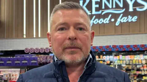 Kenny Bradley stands in his supermarket, behind him in the far distance fridges with cheese in them can be seen, to his right are bottles of sports drinks and also canned goods. Kenny has short grey hair and a short grey beard, he is wearing a dark blue padded jacket with a small Norwegian flag on its left breast.
