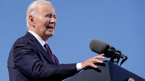 President Joe Biden speaking at a lectern, wearing a suit with a red tie