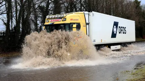 Reuters A large yellow and white lorry is driving into a flood. The water is covering the can and the road. 