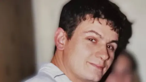 A man with short dark hair turns to the camera. He is wearing a white shirt. 