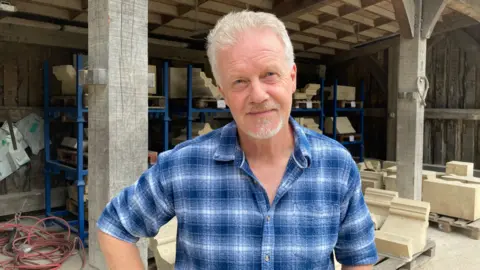 BBC/Jack Hadaway-Weller A man surrounded by carved architectural stone components stored on shelving and pallets. The workshop features timber beams, stone masonry pieces, and equipment, highlighting a traditional craftsmanship environment.
