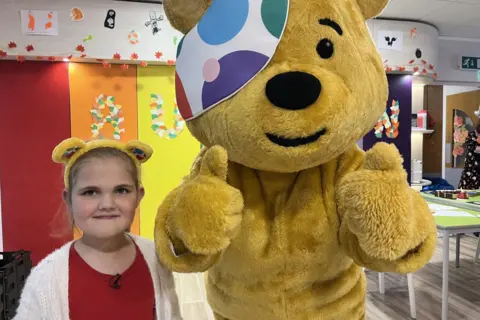 A young girl stands to the right of a life-sized Pudsey Bear. She is smiling, with dark eyes and blonde hair. She is wearing Pudsey ears. Pudsey has both thumbs up at the camera. The background is colourful, with red, orange, and yellow blocks and green tables.