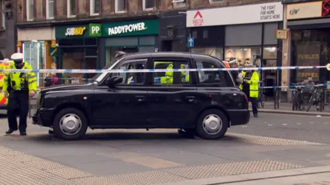 BBC Police officers in yellow coats stand beside a black cab  behind police tape