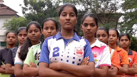 The BBC nine girls are standing in a V formation of the Kabaddi Club, crossing their arms, which was looking at the camera. Meena is in front with four girls everywhere, which is out of it. They are wearing brightly colored sports shirts.