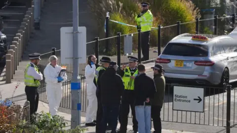 Police officers standing outside railway station entrance. There is crime scene tape in the background 