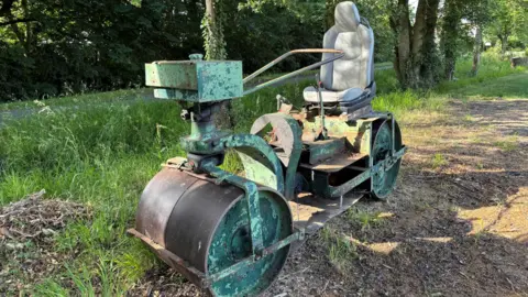 Luke Deal/BBC The club's vintage green cricket pitch roller parked under some trees on a grass verge. It has two big metal wheels at the front and back, along with a comfy seat and steering column.