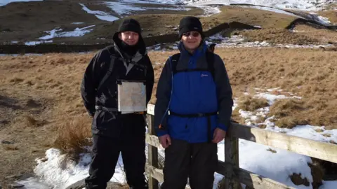 Family David Bown and his dad Steve stand at the foot of a large hill. There is snow on the ground and they are both wearing woolly hats, waterproof coats and walking trousers. 