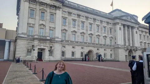 Contributed A wider view of Belinda Bradley standing outside Buckingham Palace. The building has multiple storeys with dozens of windows. She is smiling at the camera.