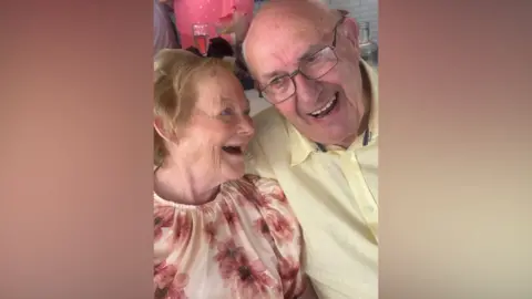 An elderly couple embrace. She has light hair and a floral top. He has grey short hair and glasses. He wears a yellow shirt. They are both smiling