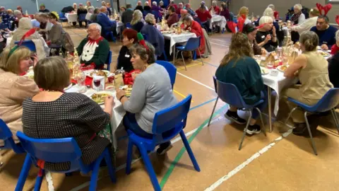Margaret Bull A number of people eating dinner at Tonbridge Baptist Church in Tonbridge on Christmas Day, 2024. People can be seen at long tables with blue plastic chairs.