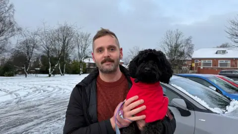 A man holds a black dog in his arms in a car park with snow covering the surface. He has a short black beard and moustache with short dark brown hair on his head. He wears a dark red jumper with a black jacket over it. The dog looks into the camera and wears a red coat over its black fur.